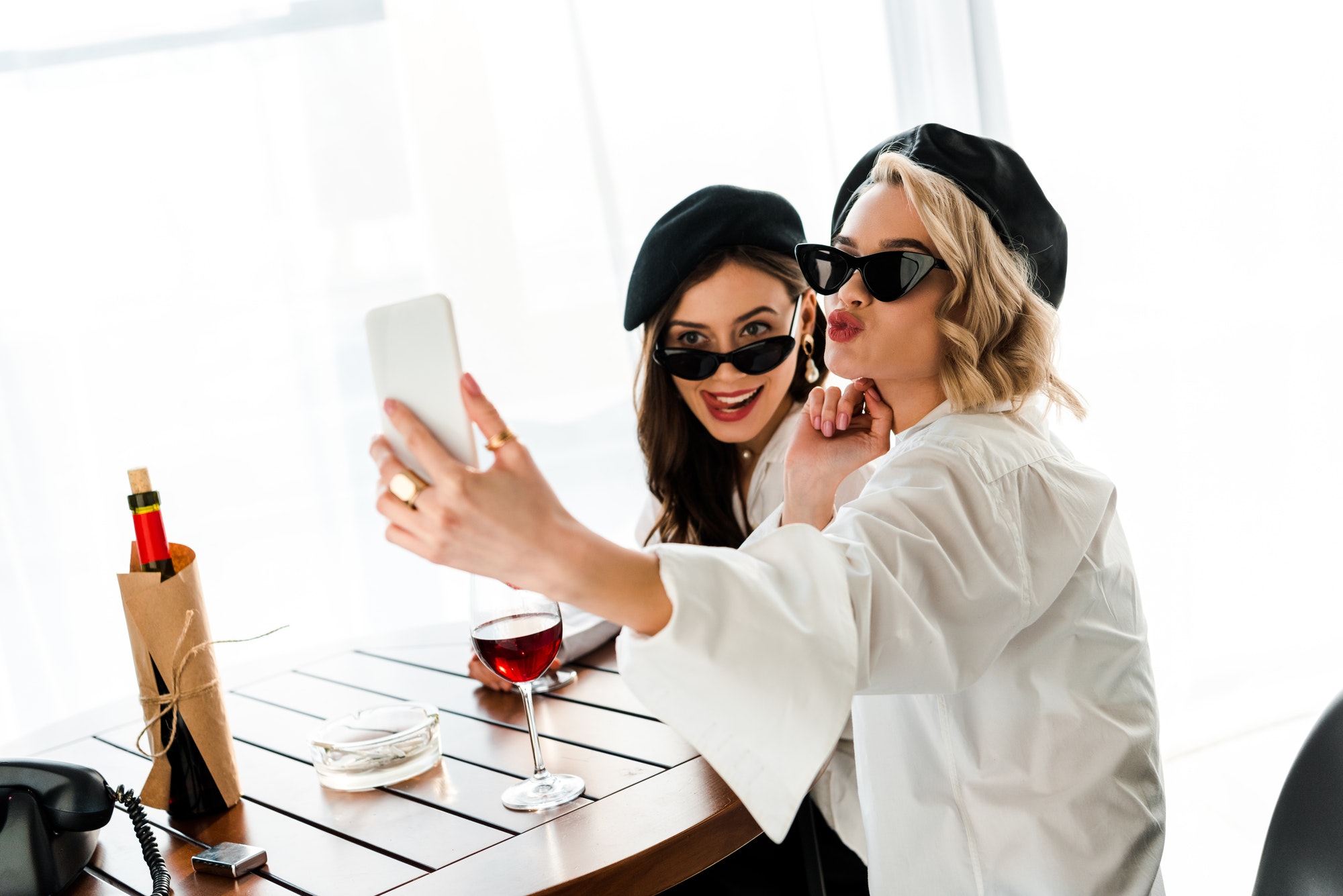 elegant brunette and blonde women in black berets and sunglasses drinking red wine and taking selfie