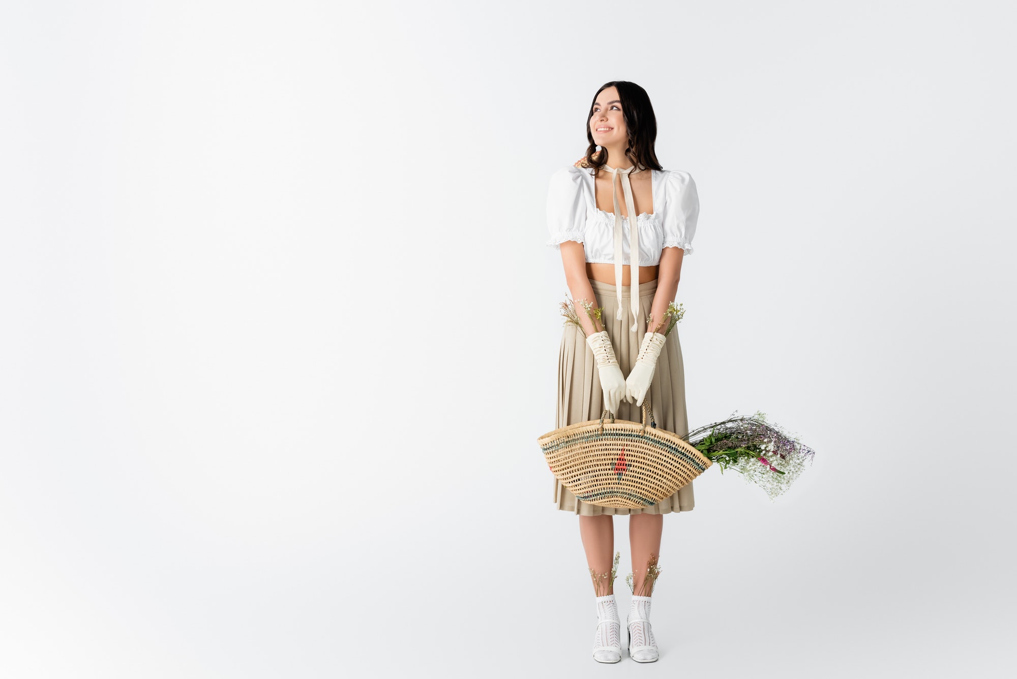 full length of joyful young woman in spring outfit holding straw bag with flowers on white